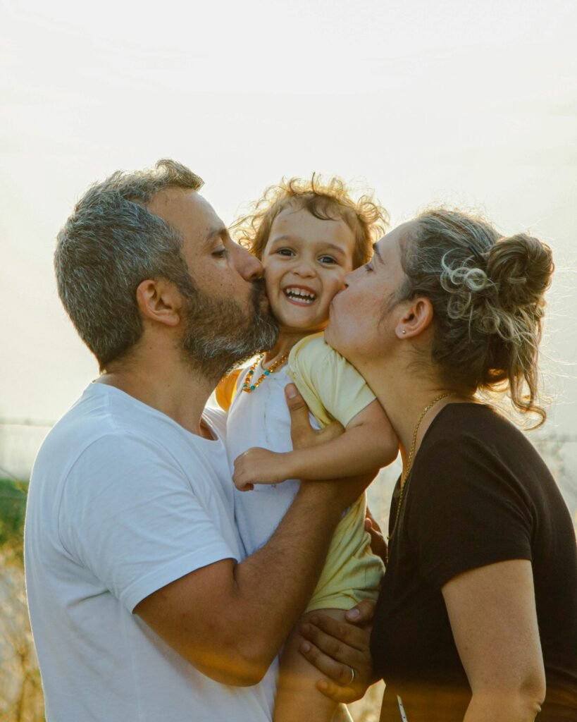 A joyful family moment with parents kissing their smiling child outdoors at sunset.